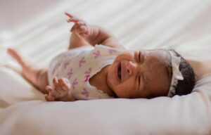 A close-up of a crying baby wearing a headband, lying on a soft bed indoors.