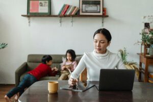 Mother multitasking with laptop and stylus alongside her children playing at home.