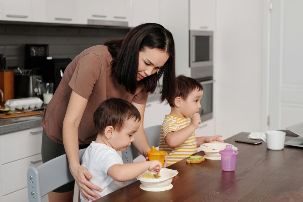 A mother helps her toddlers enjoy breakfast in a modern kitchen setting.