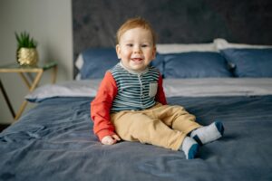 Cute toddler in striped shirt and red sleeves sits smiling on a cozy bed indoors.
